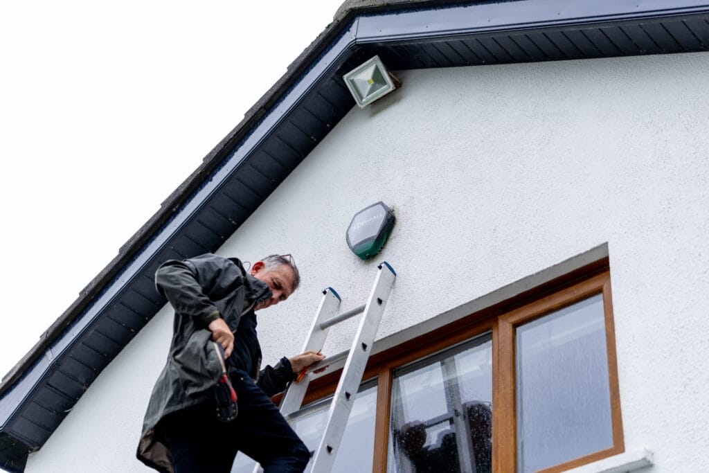 Engineer on aluminium ladder installing black and green alarm bell box on white render